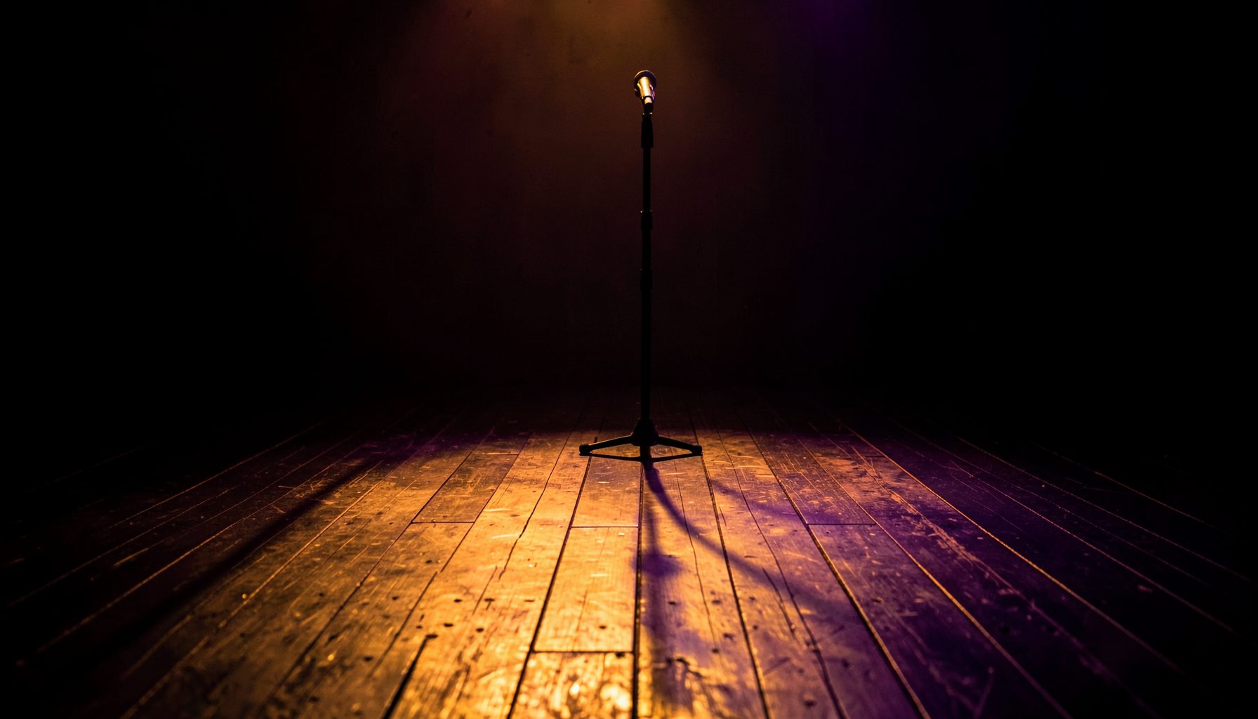 Dark stage with warm amber and violet lights casting long shadows across worn wooden floorboards, a microphone stand silhouetted against a moody backdrop