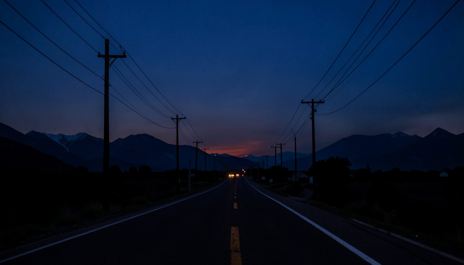 A dimly lit rural highway stretching toward distant mountains under a deep indigo dusk sky, with telephone poles lining the road and a faint warm glow on the horizon.